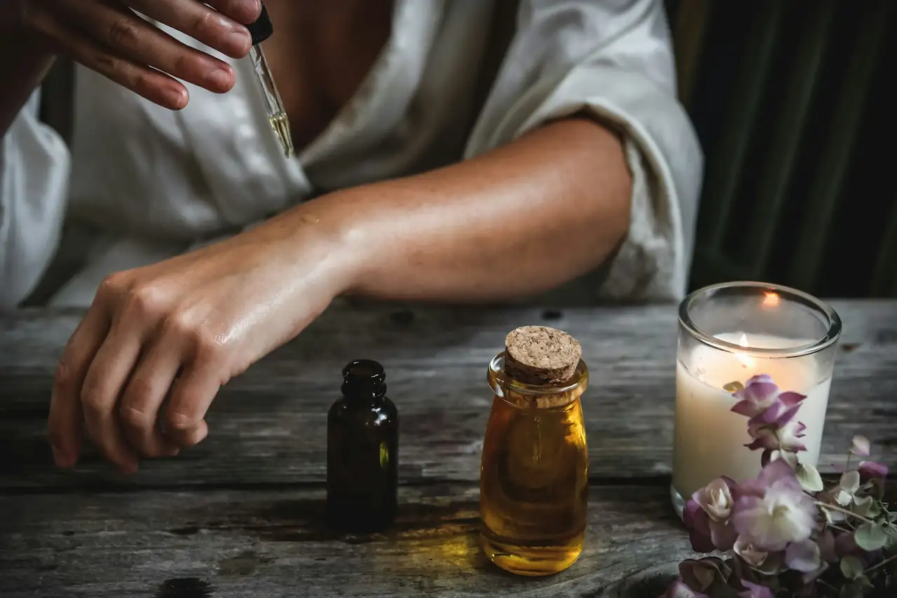 A dropper of amber oil over a wrist, with a glass bottle and a candle on a wooden table